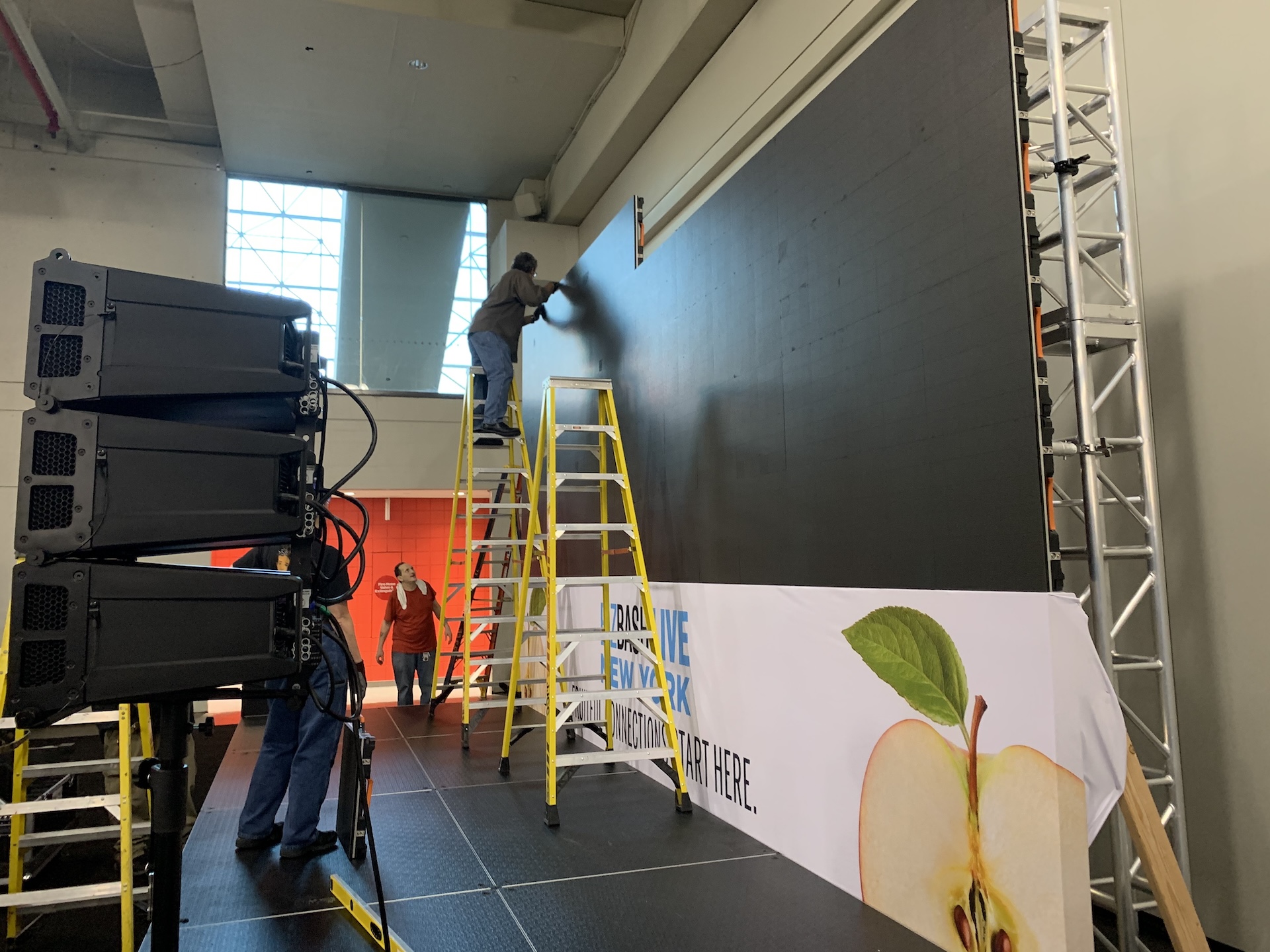 One technician on a ladder building an LED wall for an event while another technician works with a speaker system