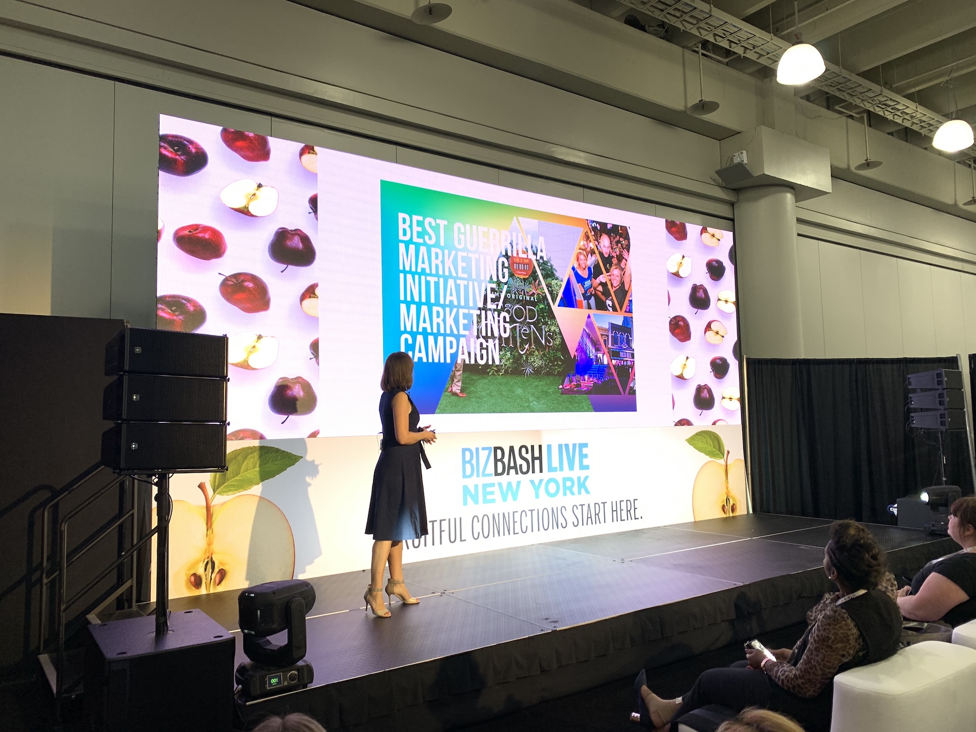 A presenter at BizBashLive New York presenting in front of a screen with bright visuals in triangle shapes