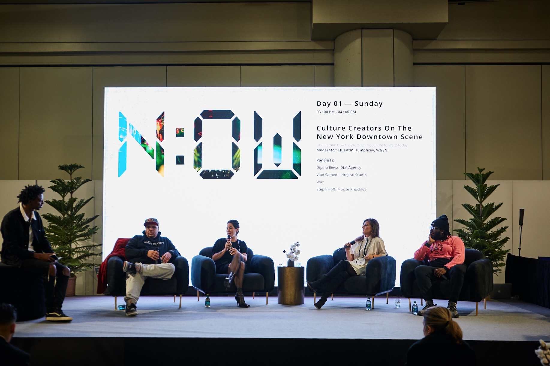 A panel in comfortable arm chairs sitting in front of a screen that reads "N:OW" in big print